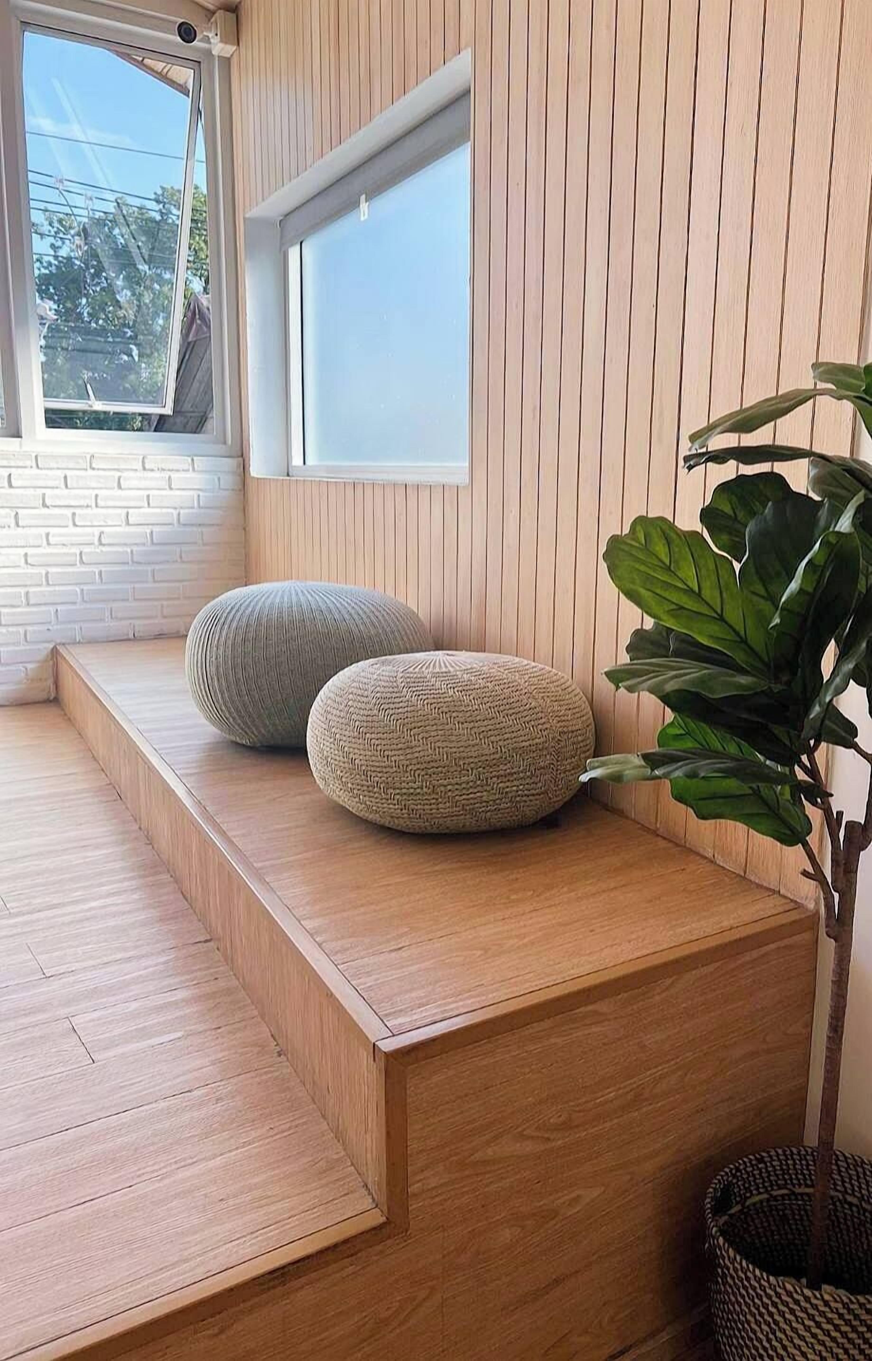 A wooden bench with two woven poufs, a large plant, and a window with blinds.