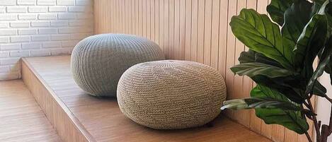Un banco de madera con dos otomanas tejidas, una planta grande y una ventana con persianas.