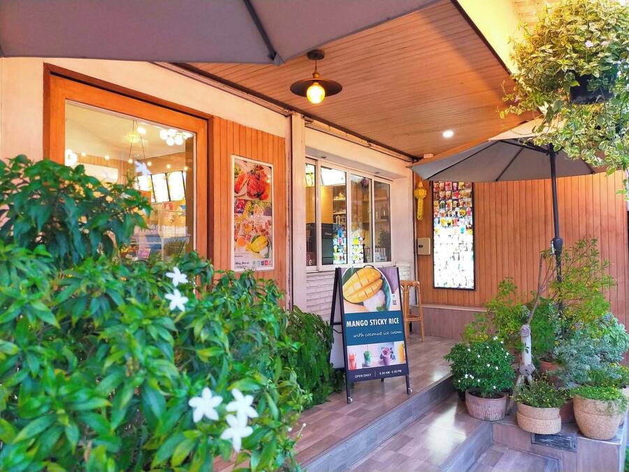 A restaurant entrance with a wooden facade, a poster advertising mango sticky rice, and potted plants.