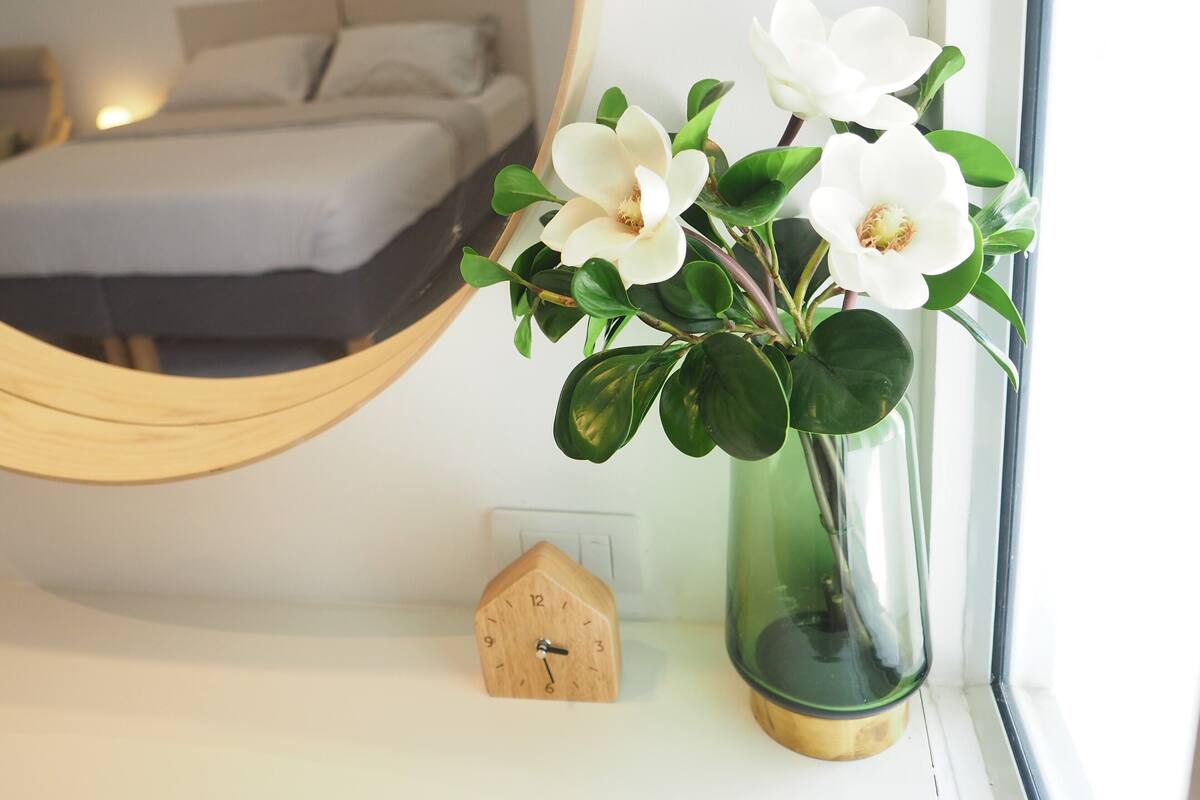 Un dormitorio con cama, una mesita de noche con un reloj, un espejo y un florero con flores.