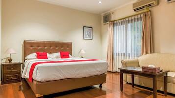 A hotel room with a large bed, a bedside table, a chair, and an air conditioning unit.