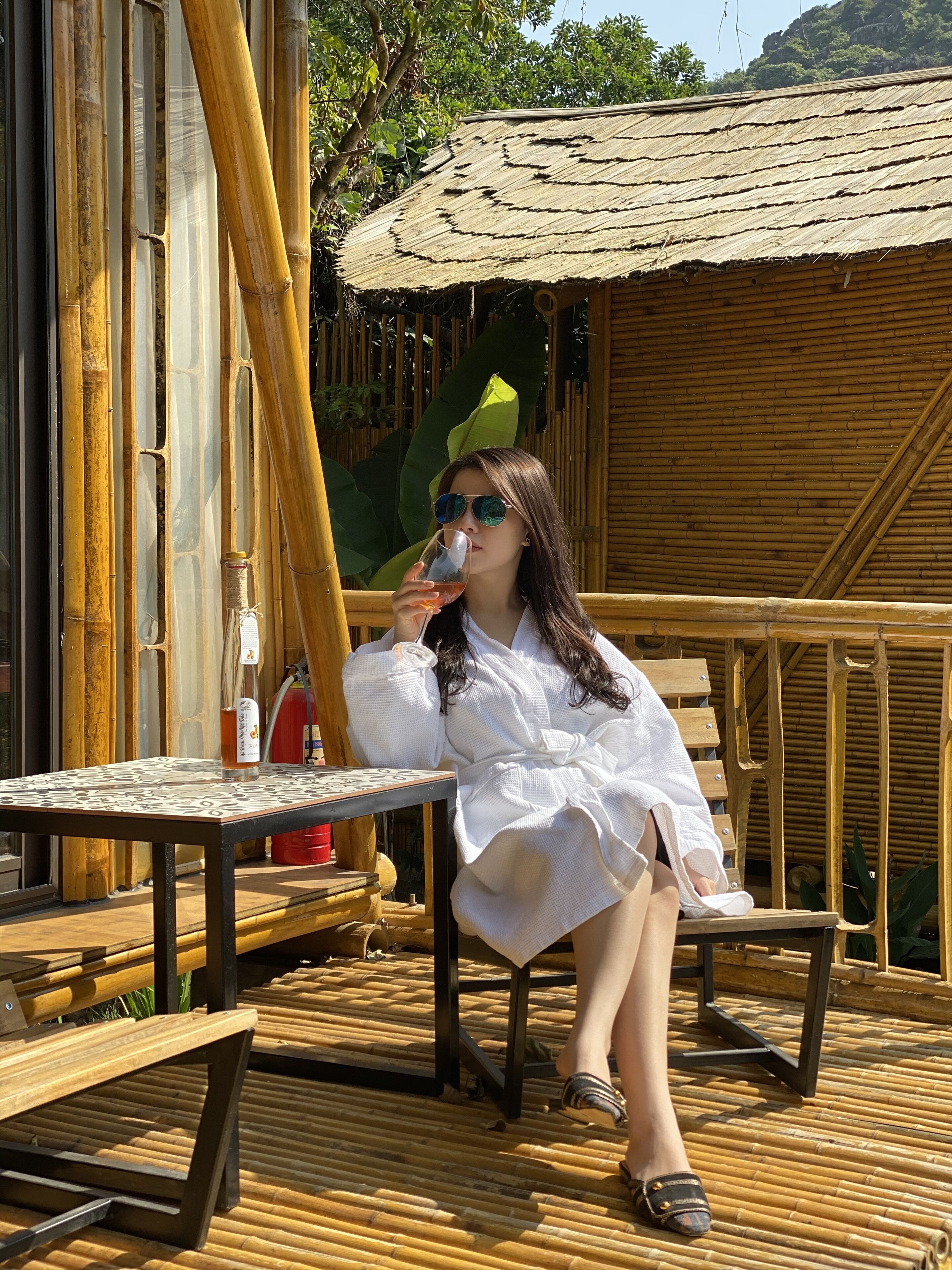 A person sitting on a wooden deck with a table and chairs, a thatched roof structure, and bamboo walls.