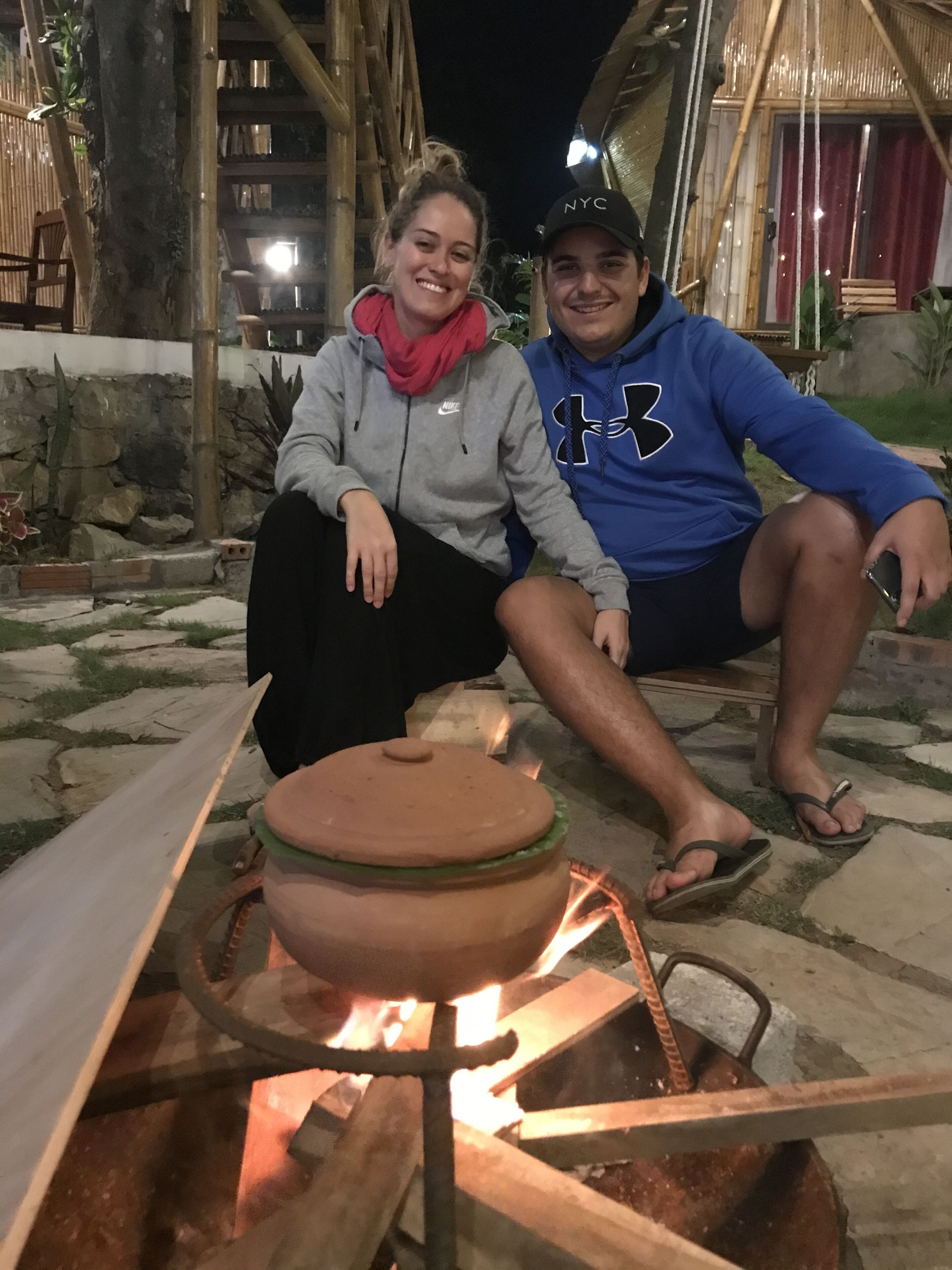 Two people sitting by a campfire with a pot on a portable stove.