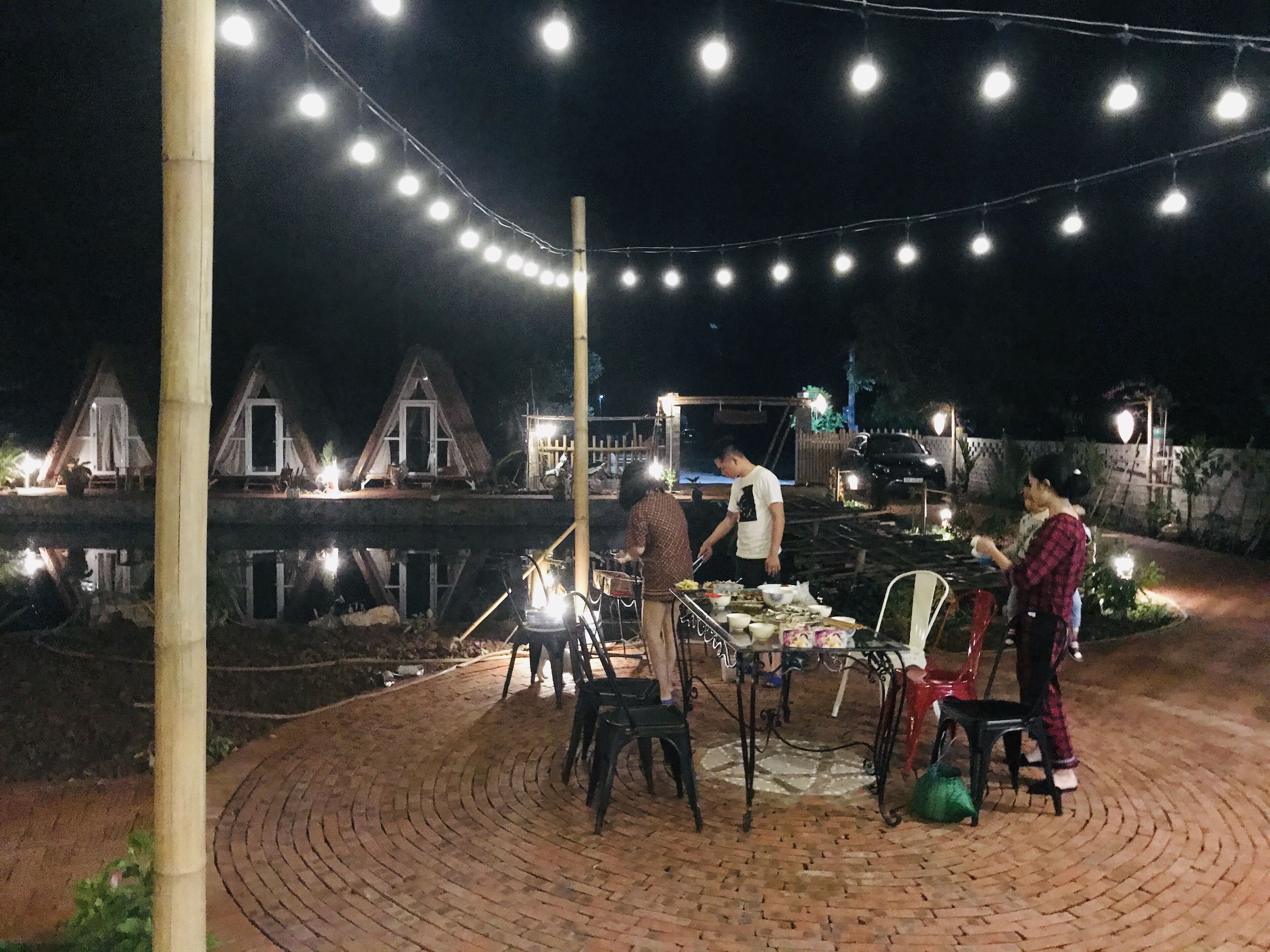 Een terras in de open lucht 's nachts, met lichtjes, een terrasvloer van baksteen en uitzicht op een waterpartij met gebouwen op de achtergrond.