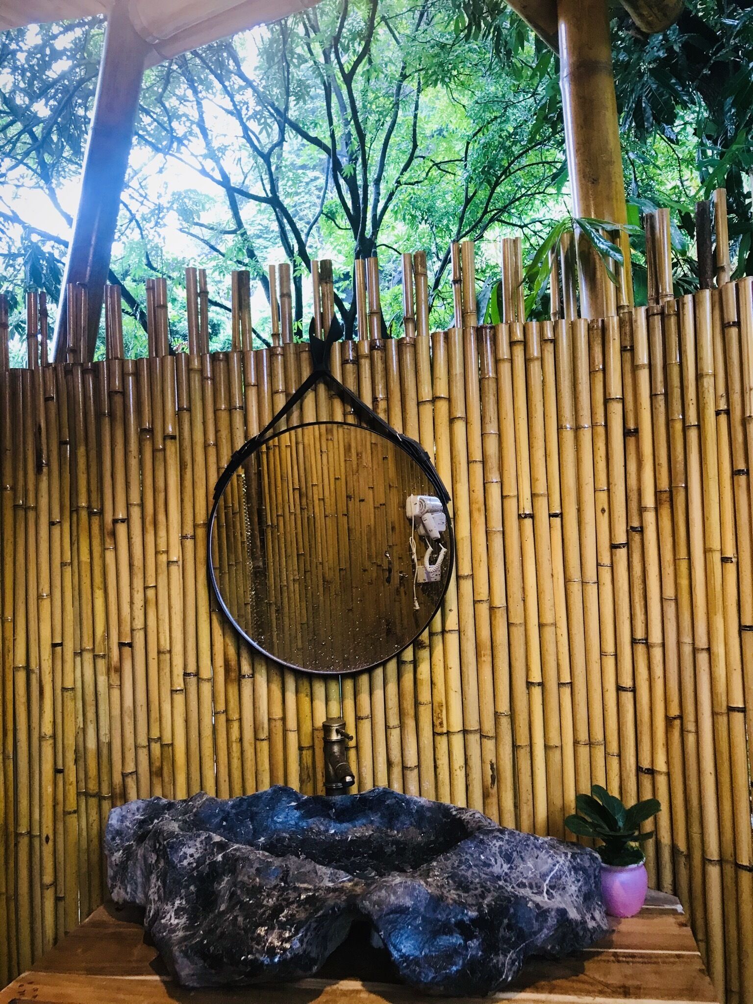 A bathroom with a bamboo wall, a round mirror, a sink with a stone basin, and a small potted plant.
