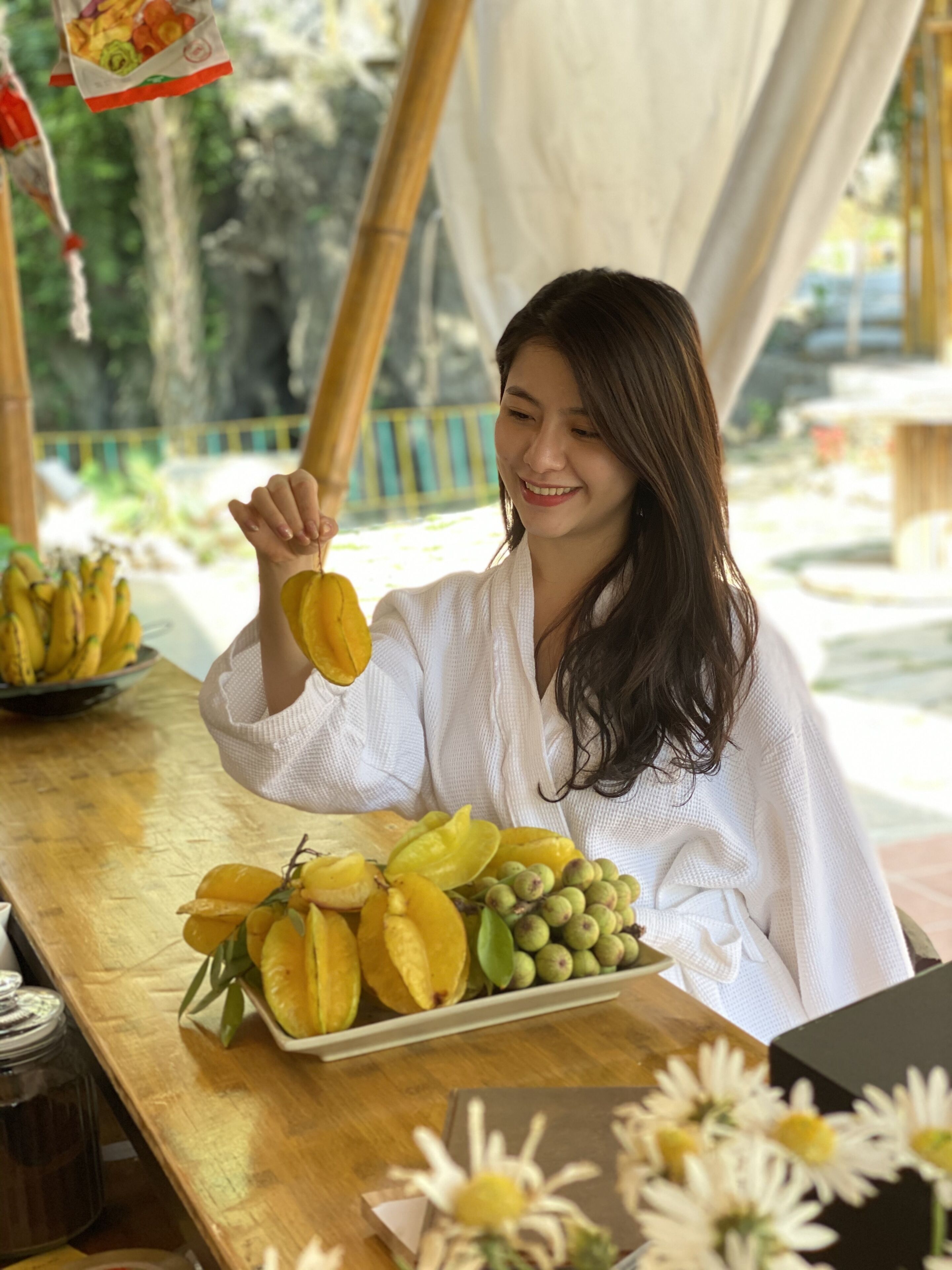 A person holding a fruit at a wooden table with a tray of fruits and flowers.