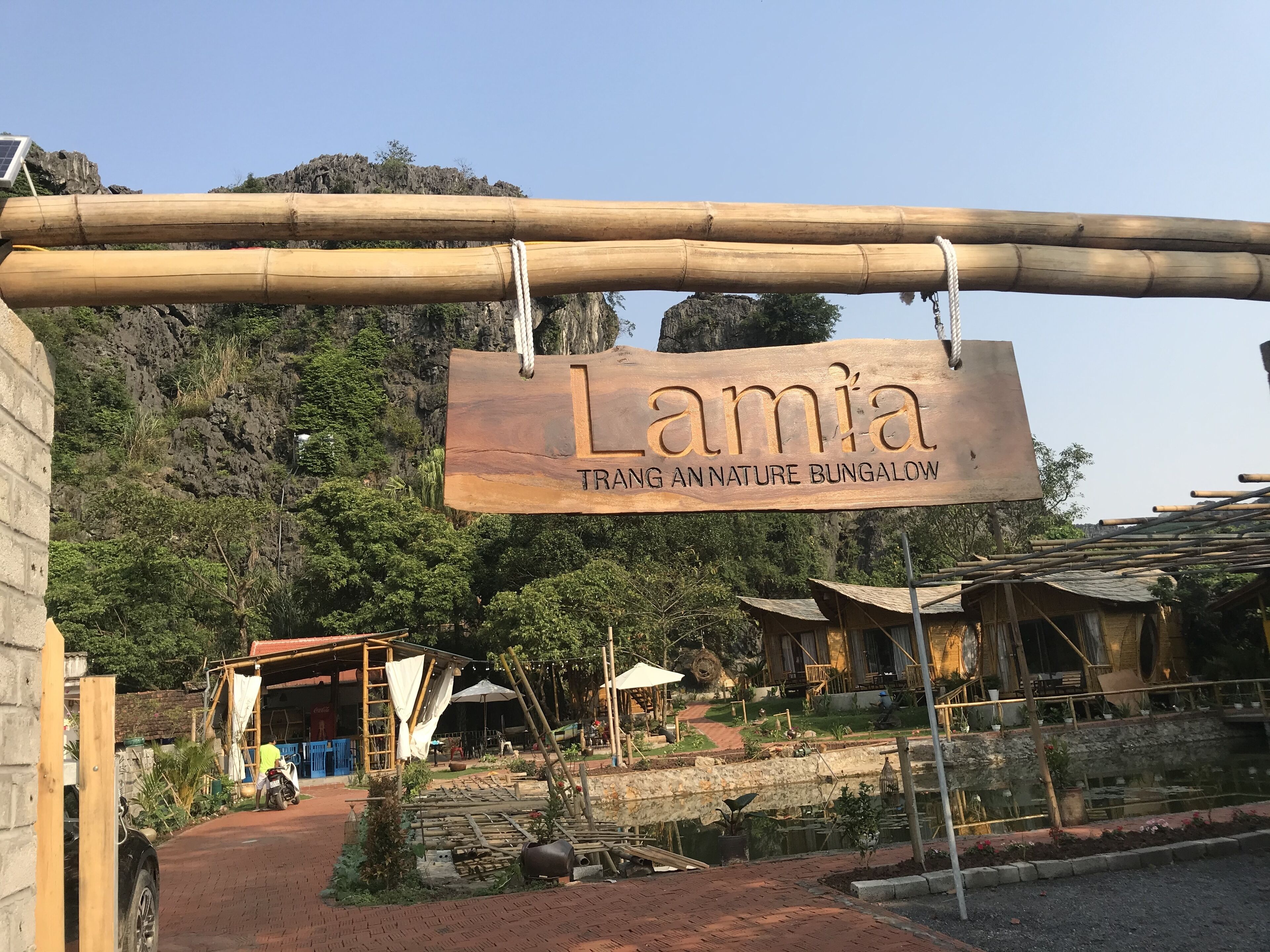 Un letrero de madera indica 'Lamia Trano Nature Bungalow', en un entorno rústico que incluye construcciones de bambú, un jardín y un sendero.