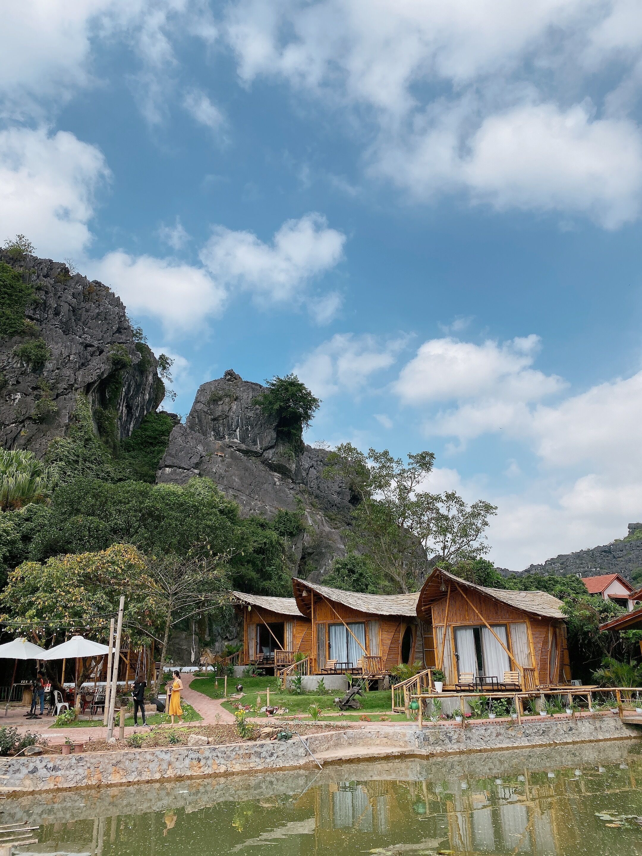 Uma cabana rústica de madeira com um caminho de pedra que a leva, cercada por vegetação exuberante e um penhasco rochoso.