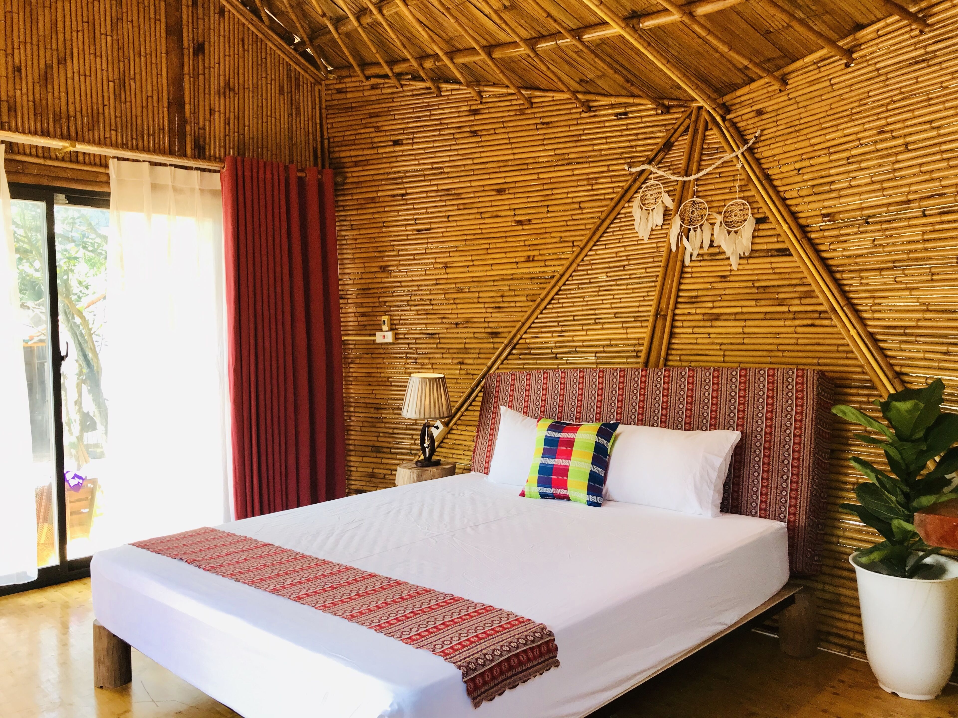 A room with a bamboo ceiling, a bed with a white mattress and red and white patterned bedspread, a colorful pillow, and a bedside lamp.