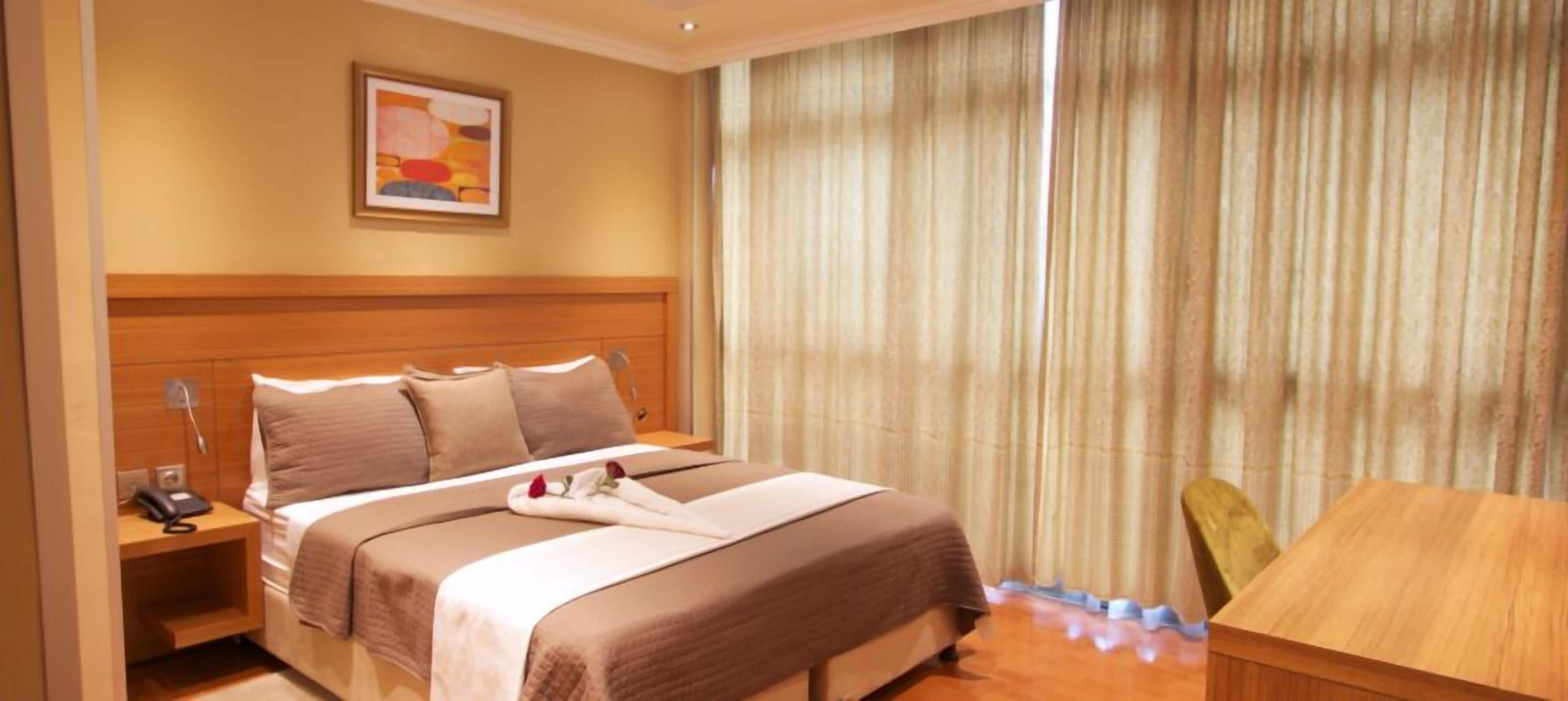 Une chambre d’hôtel avec un grand lit, une tête de lit en bois, une table de chevet avec un téléphone, un tableau au mur et une vue sur un rideau.