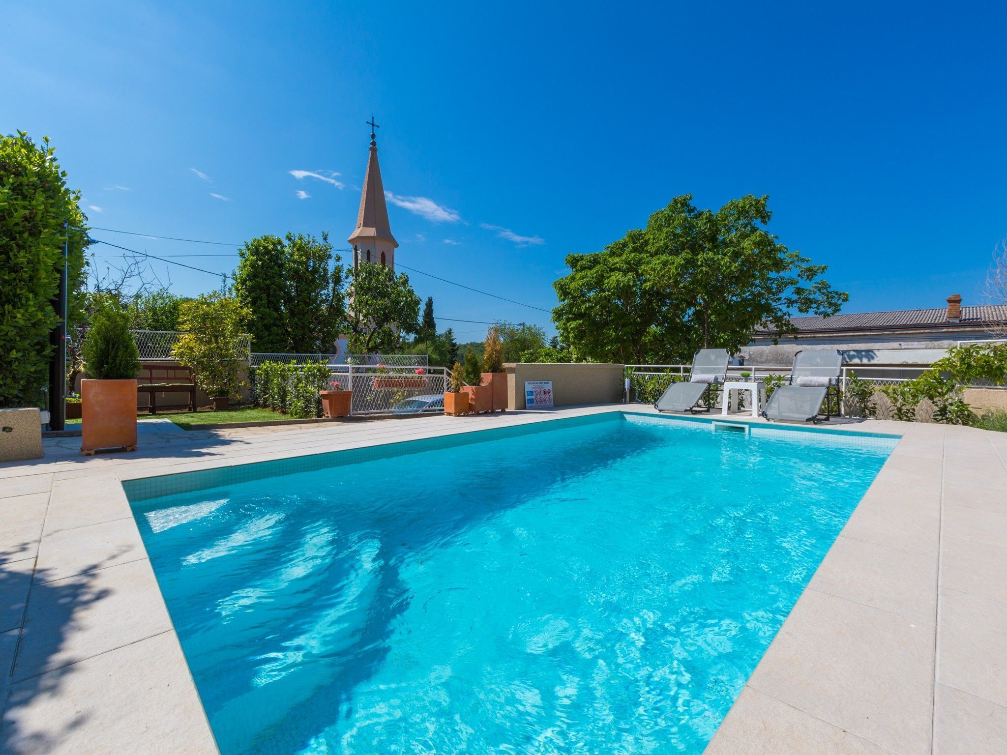 Una piscina con lettini, una recinzione e una guglia di una chiesa sullo sfondo.