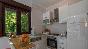A modern kitchen with white cabinetry, a built-in oven, a stove, and a microwave. There is a window with a view of greenery outside, and a wooden cutting board with oranges on the counter. - House Lara (Jadranovo)