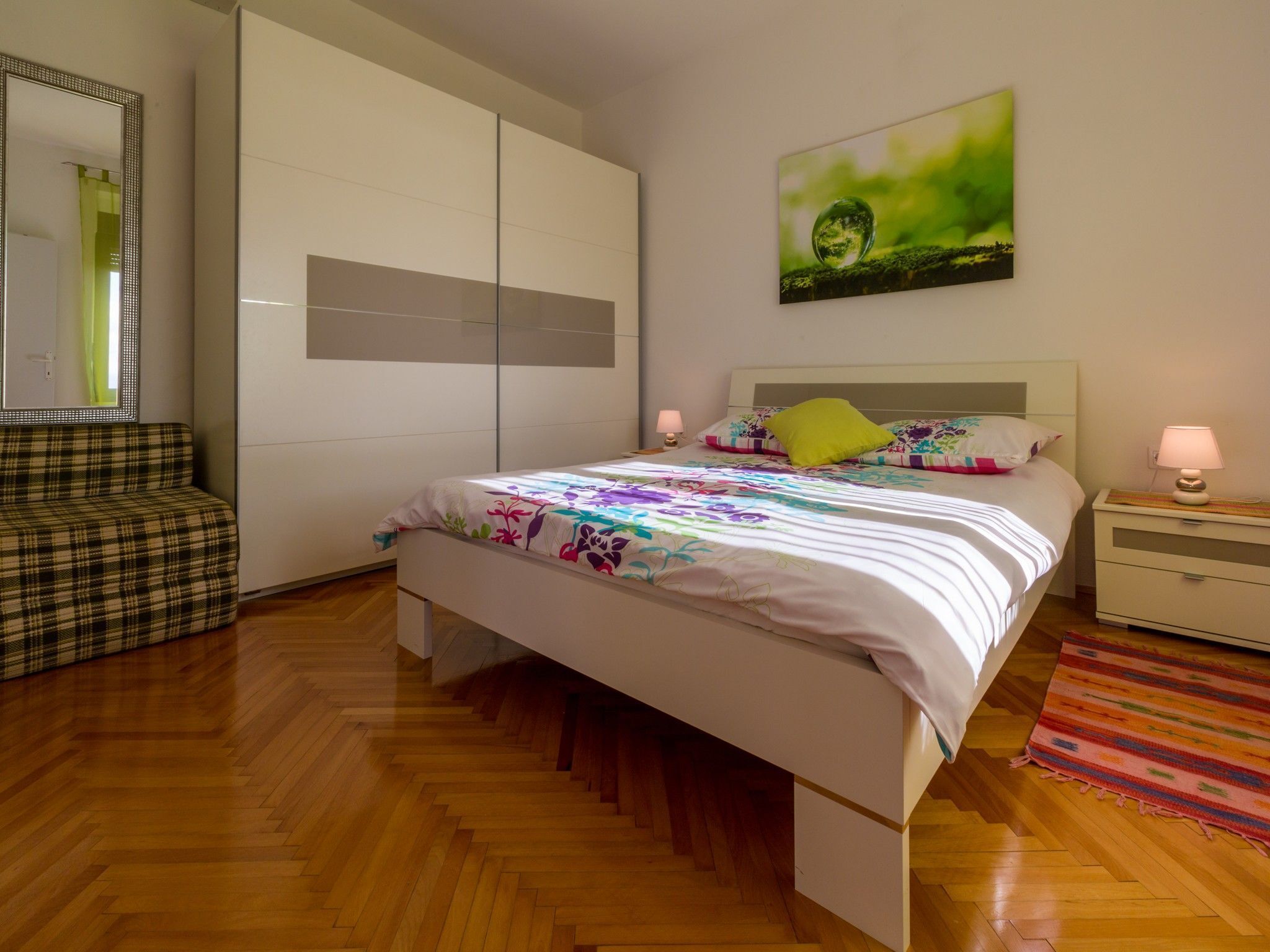 A bedroom with a bed, a nightstand, a lamp, a mirror, a sofa, and a painting on the wall.
