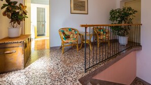 A spacious room with a tiled floor, a wooden chest, a dining table with chairs, a staircase with a railing, and a potted plant.