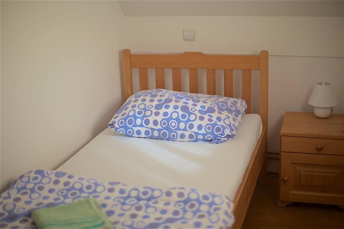 A single bed with a wooden headboard, a bedside table with a lamp, and a small drawer unit.