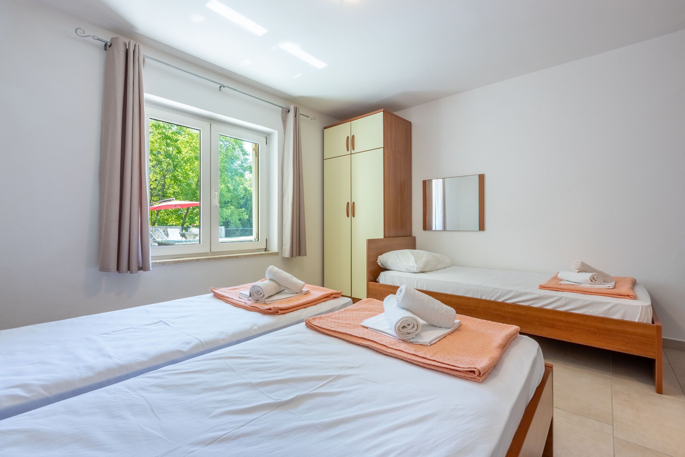 A hotel room with two single beds, a wardrobe, a window with curtains, and a view of greenery outside.