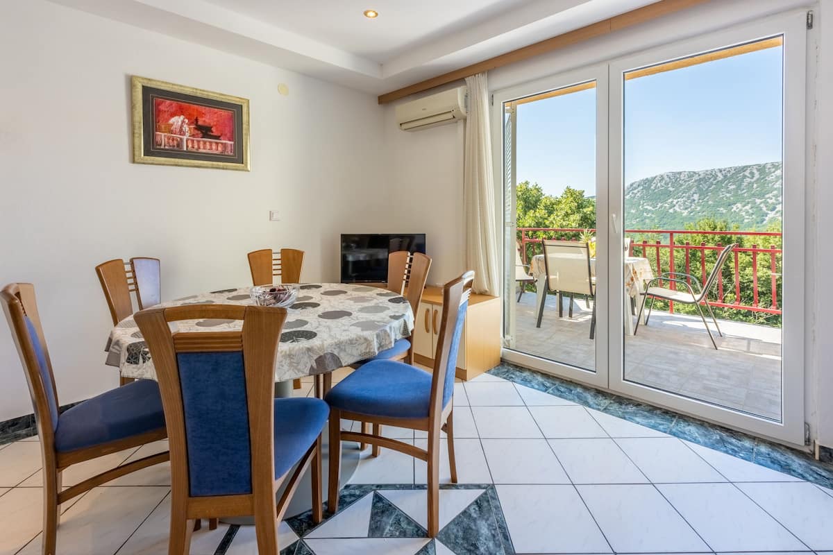 A dining area with a table and chairs, a TV, and a balcony with a view of the mountains.
