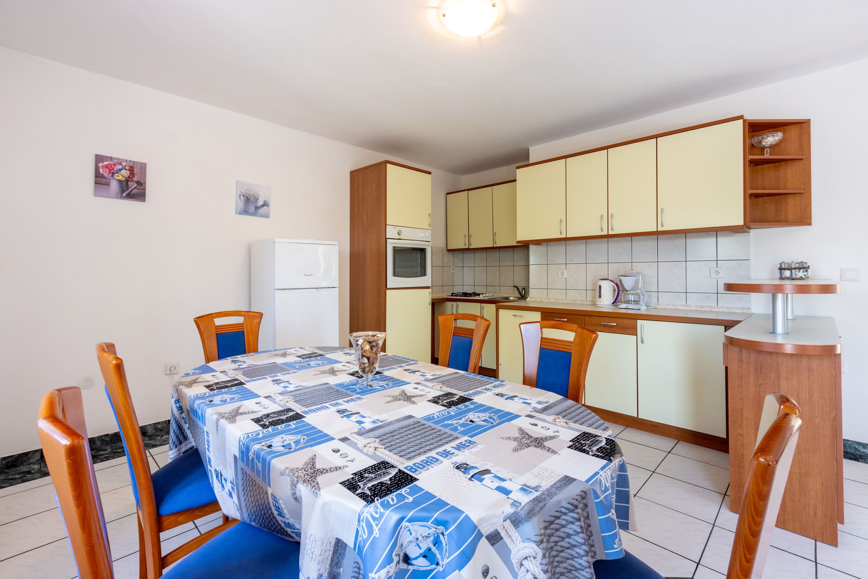 A compact kitchen with a dining area, featuring a table with a blue and white tablecloth, wooden chairs, and a small refrigerator.