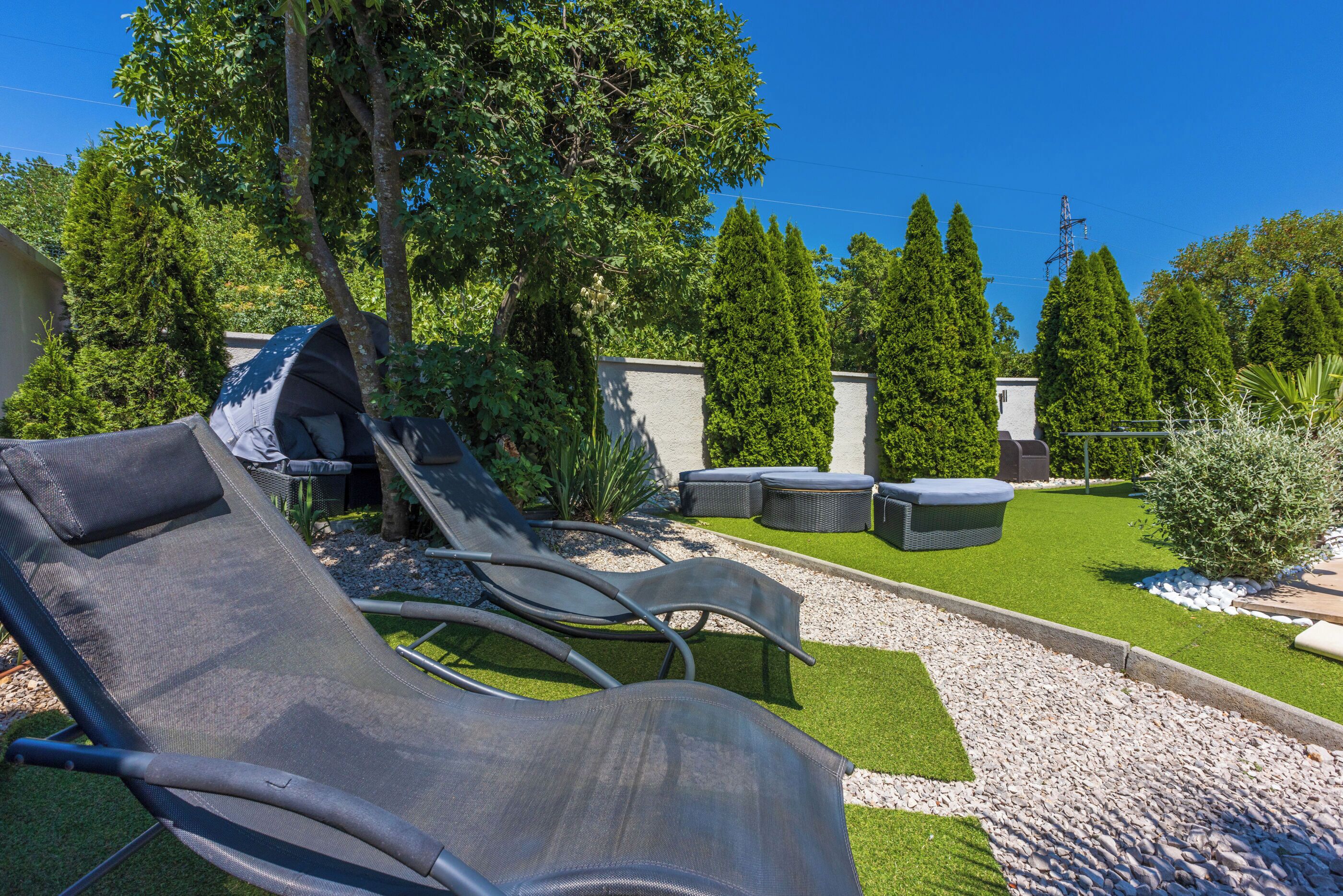 Ein Garten mit Sitzgelegenheiten im Freien, darunter Lounge-Stühle und ein kleiner Tisch, umgeben von viel Grün und blauem Himmel.