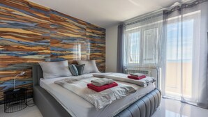 A modern bedroom with a large bed, a wooden wall panel, a bedside table, and a window with blinds. - House Moderna 2 (Crikvenica)