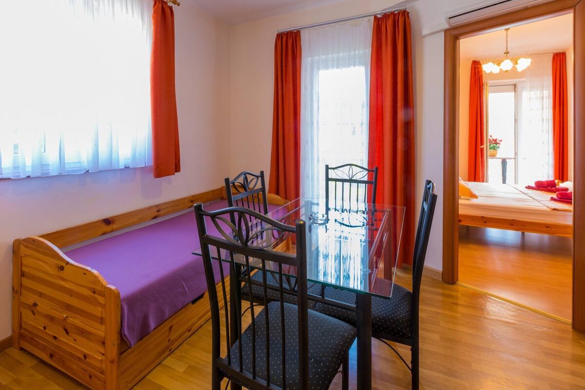 Ein Zimmer mit einem hölzernen Bett, einem Glastisch mit Stühlen und einem Fenster mit roten Vorhängen.