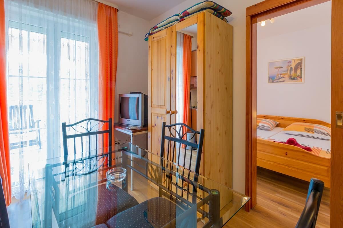 Ein Zimmer mit einem Glastisch, Stühlen, einem Fernseher, einem Bett und einem Holzschrank.