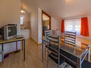 Ein kompakter Wohnbereich mit einer Küchenzeile, einem Essbereich mit Glastisch und Stühlen, einem Fernseher auf einem Tisch und einem Fenster mit orangefarbenen Vorhängen.