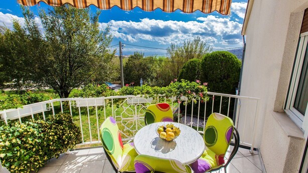 Un balcon avec une balustrade blanche, une table ronde sur laquelle se trouve un saladier rempli de fruits, et des chaises aux couleurs vives.