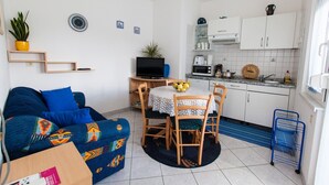Un espace de vie compact comprenant un canapé, une table à manger, un téléviseur, un coin cuisine avec des placards et des appareils électroménagers, ainsi qu’une petite plante en pot.