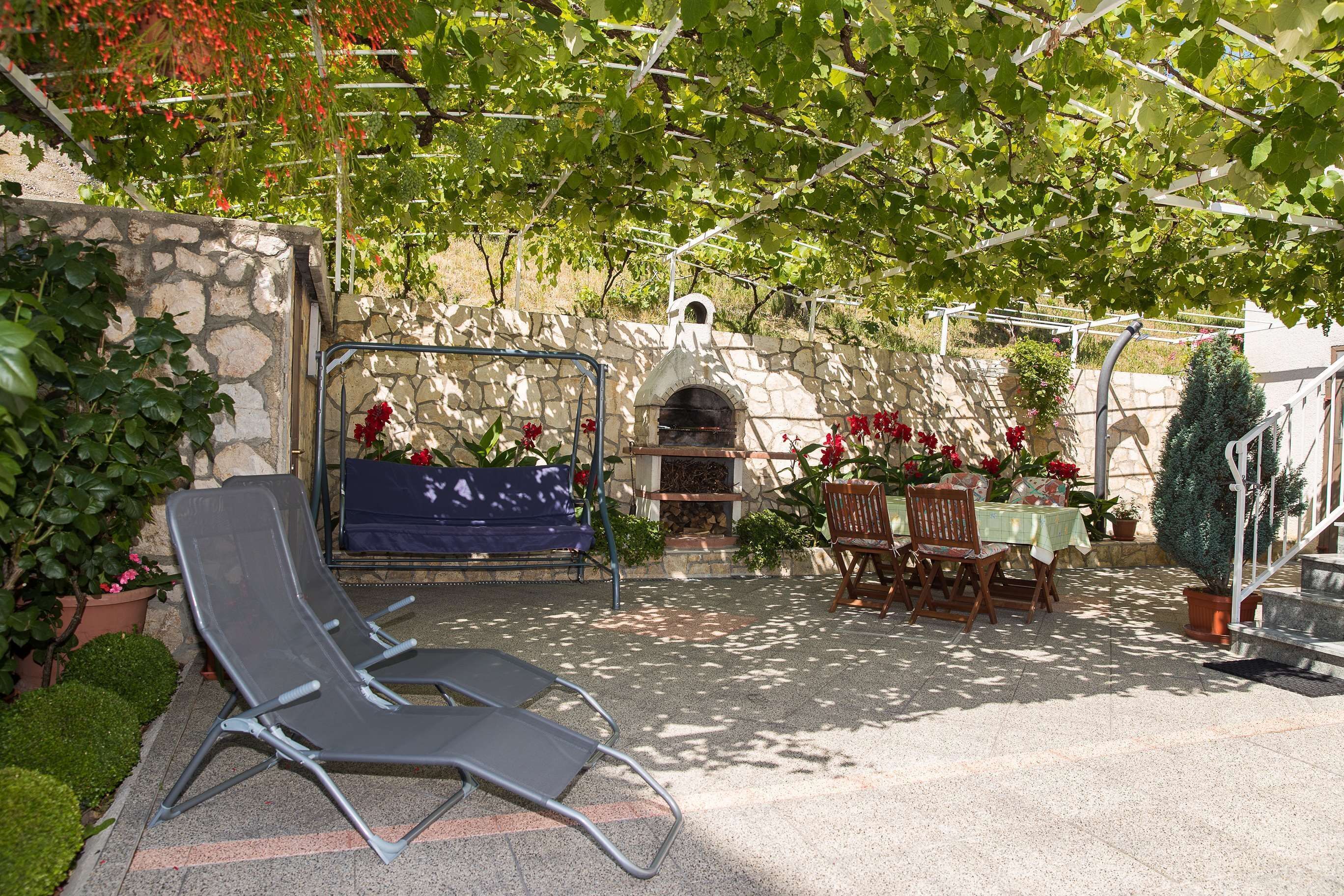 Un muro de piedra con un horno de pizza integrado, una zona de estar con mesa y sillas, y una pérgola cubierta de plantas trepadoras.