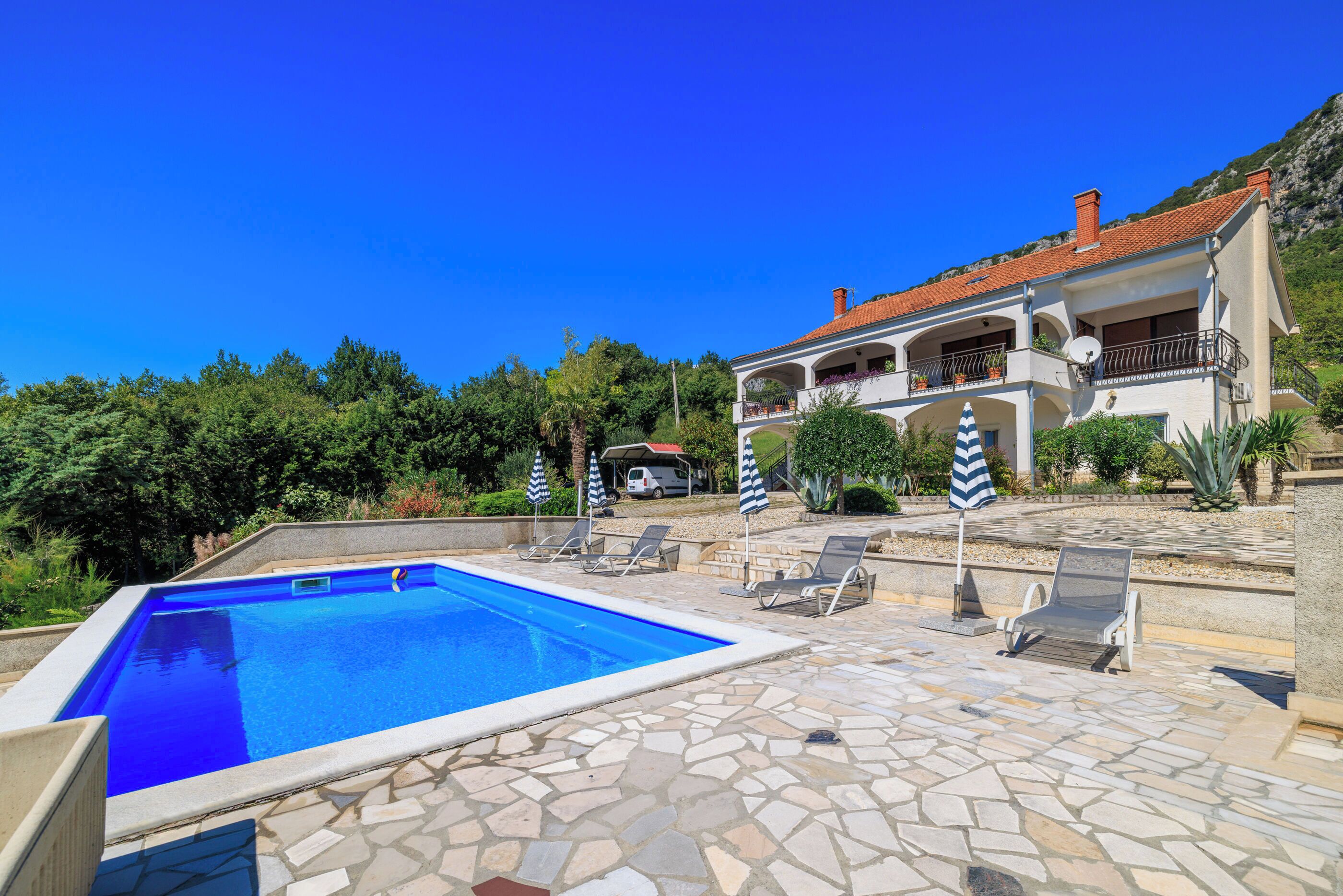 Une piscine avec des chaises longues et des parasols, un bâtiment blanc doté de balcons, et un paysage montagneux en arrière-plan.