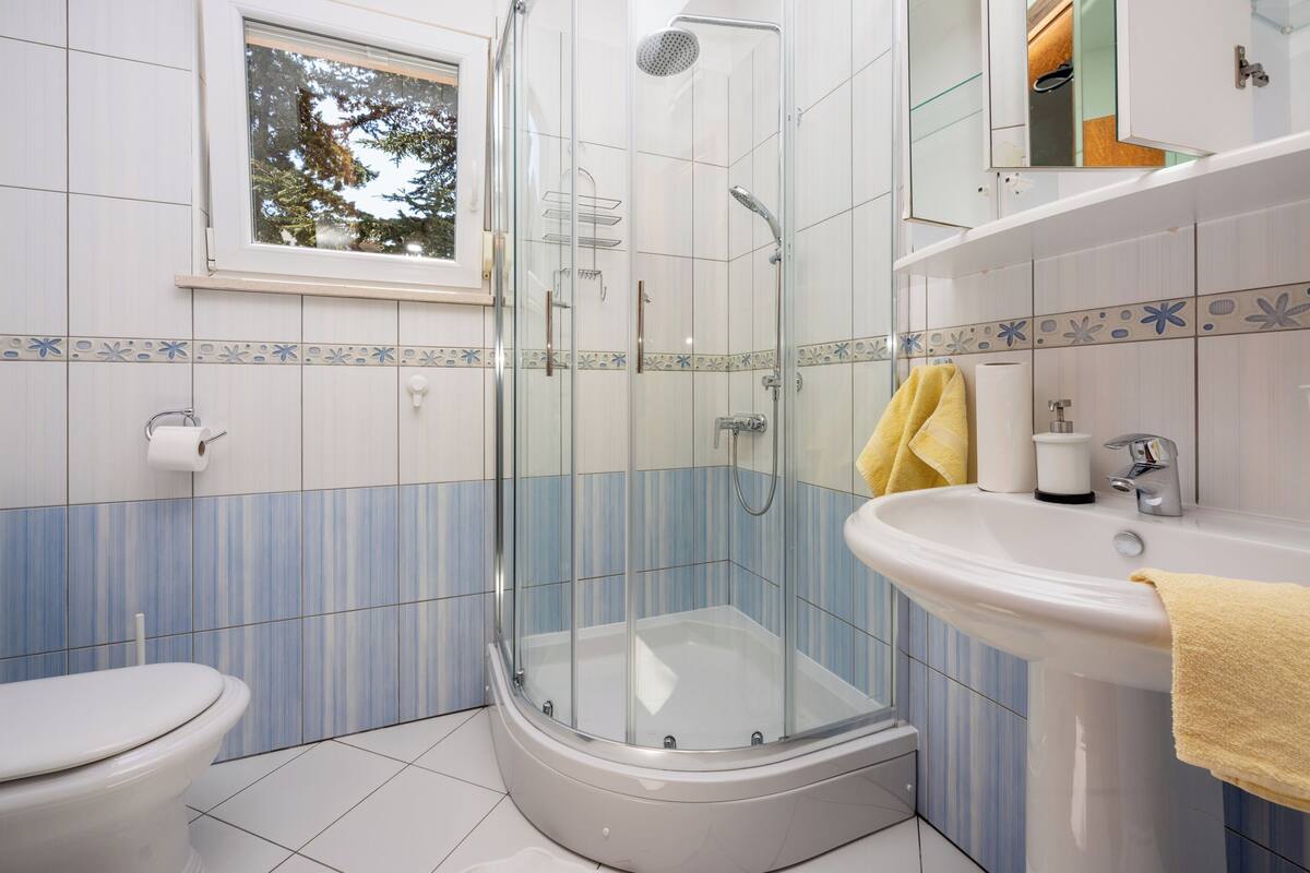 A bathroom with a glass-enclosed shower, a toilet, a sink, and a window with a view of trees.