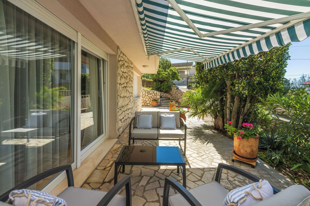 An outdoor patio with a striped awning, stone flooring, and a seating area with a table and chairs.