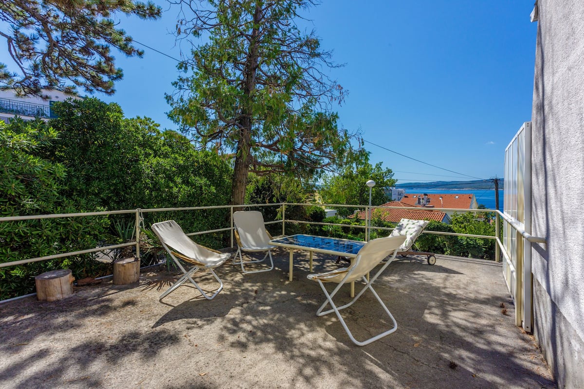 An outdoor patio with two lounge chairs and a table, surrounded by greenery and overlooking a coastal area.