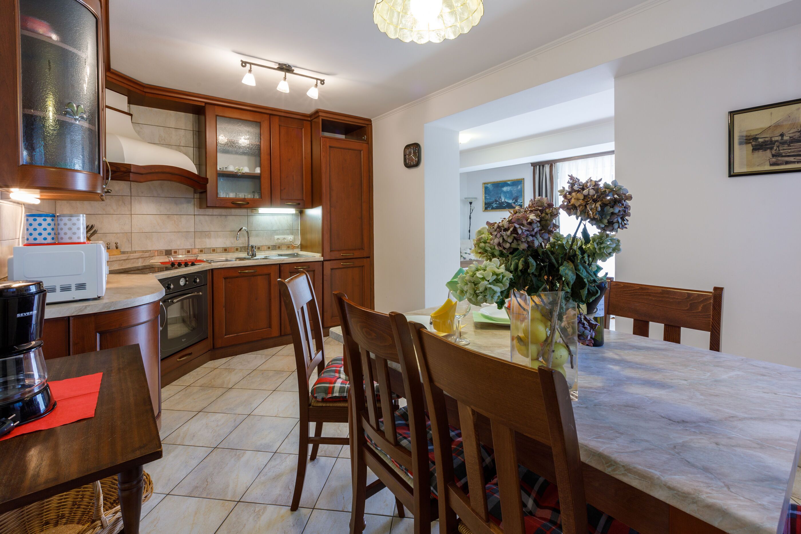 A kitchen with wooden cabinets, a dining table with chairs, a coffee maker, and a vase with flowers.