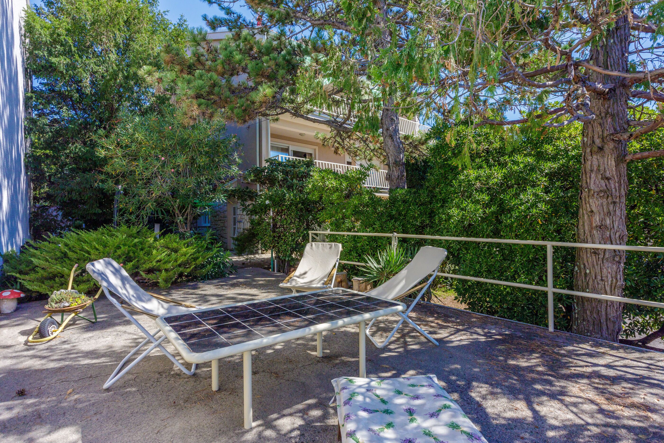 A patio with two sun loungers and a table, surrounded by dense greenery and a building in the background.