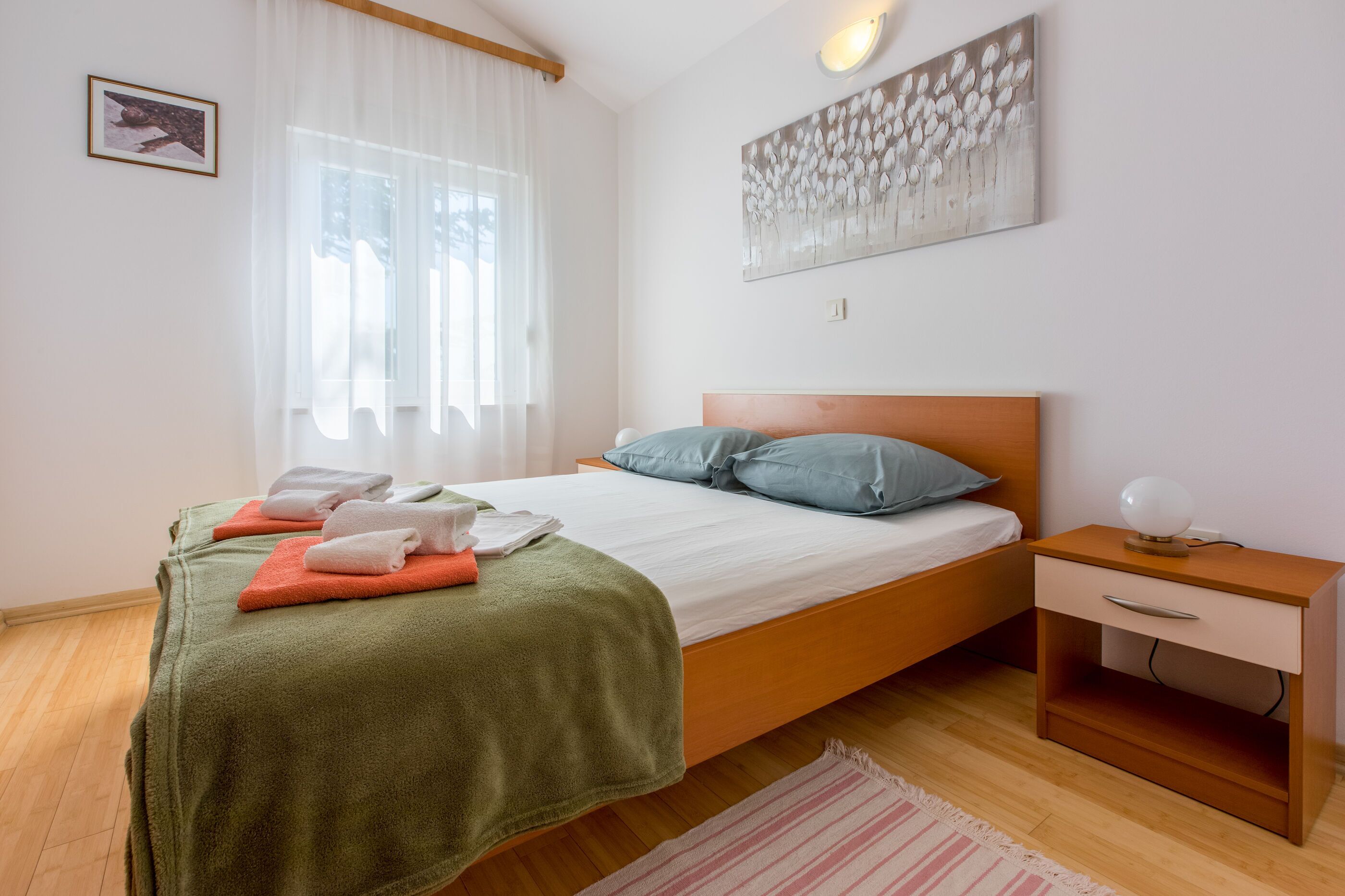 Un lit bien fait, recouvert d’une couverture verte, une table de chevet en bois avec une lampe, et un tableau accroché au mur.