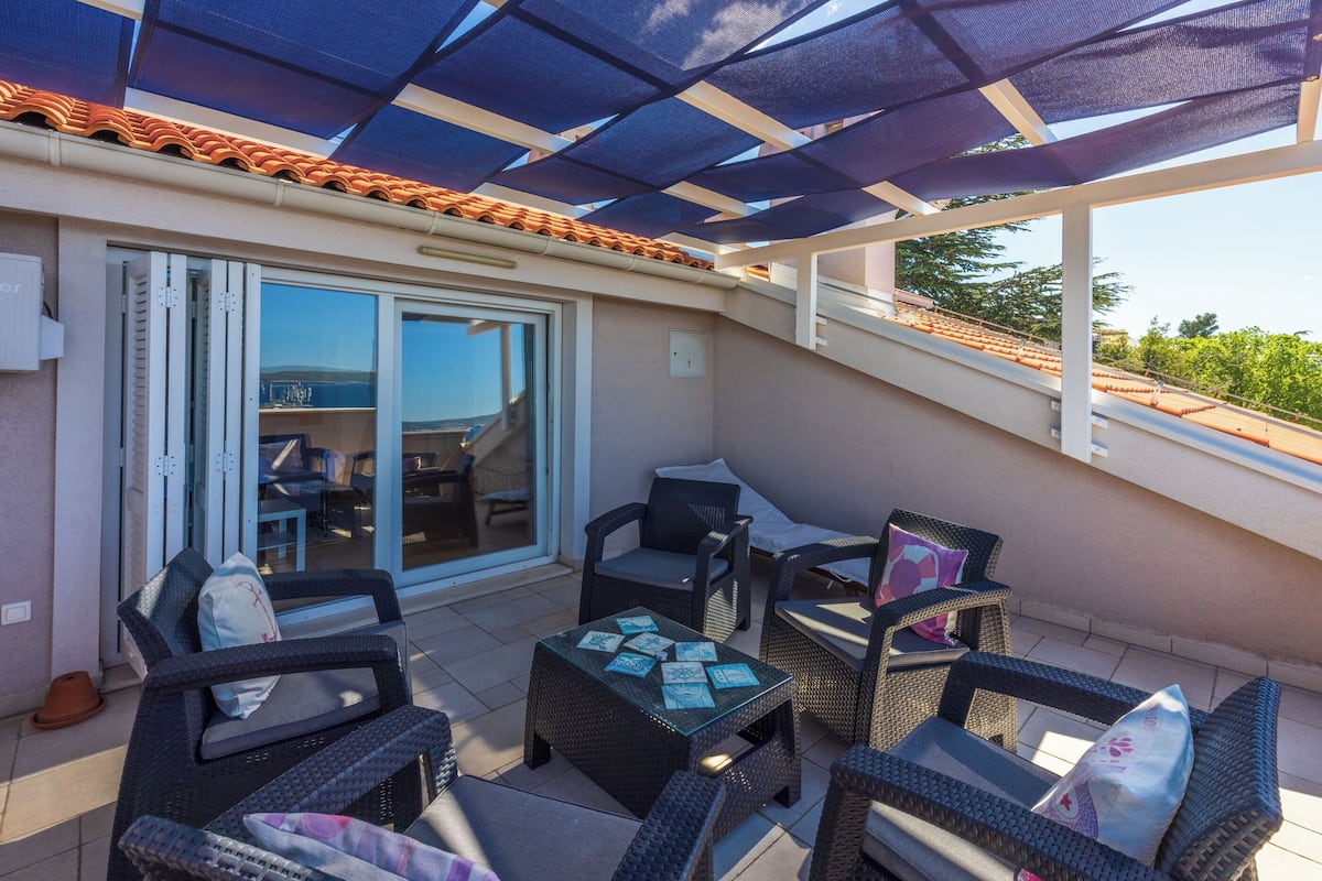 A covered outdoor seating area with wicker furniture and a glass table, overlooking a scenic landscape.