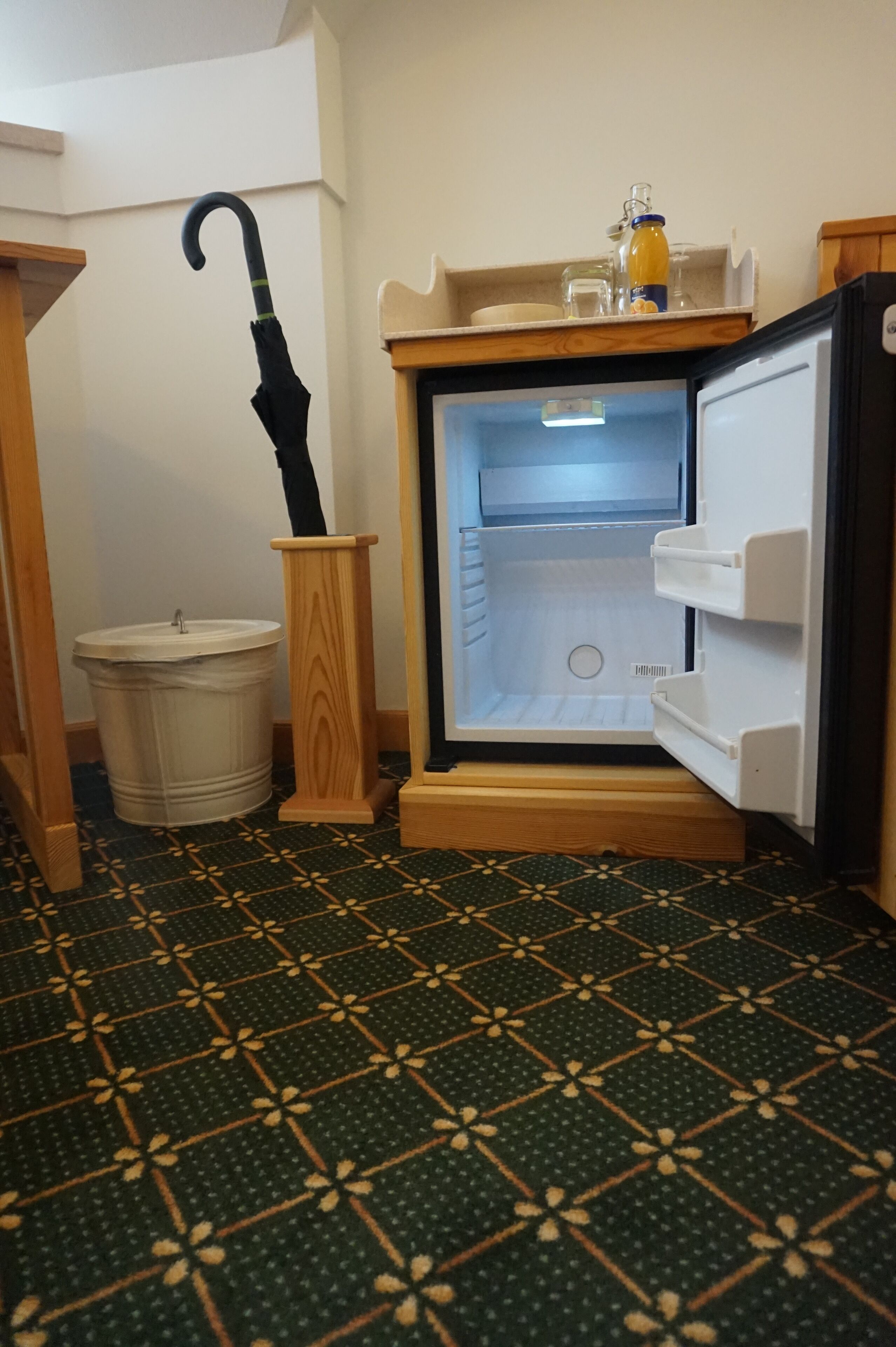 Ein Zimmer mit einem gemusterten Boden, einem hölzernen Sockel mit einem Regenschirm, einer weißen Mülltonne und einem offenen Kühlschrank.