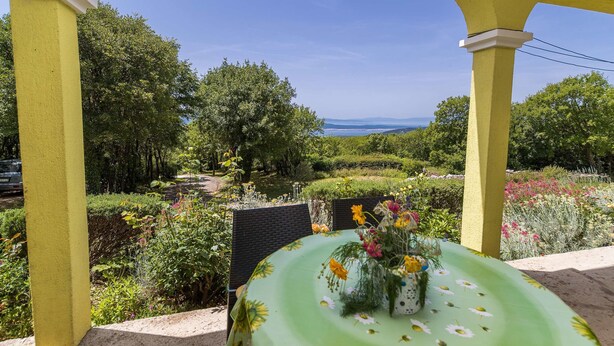 Un área de estar al aire libre con techo, con una mesa lista para dos personas, rodeada de abundante vegetación y vistas al paisaje.