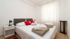 Una cama bien hecha con ropa blanca y varios almohadones, una mesita de noche con lámpara, una pequeña mesita con una taza, y piso de madera.