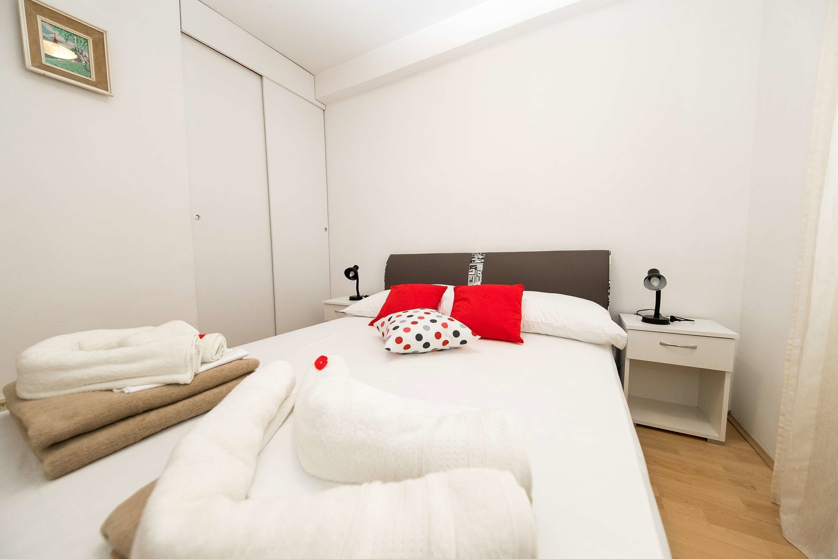 Een net opgemaakt bed met witte en bruine handdoeken, rode en stippeldeken kussens, en een nachtkastje met een lamp.