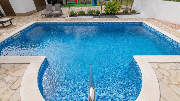 A swimming pool with a tiled edge, surrounded by stone tiles and a small garden area with plants and a bench. - House Marija (Labin)