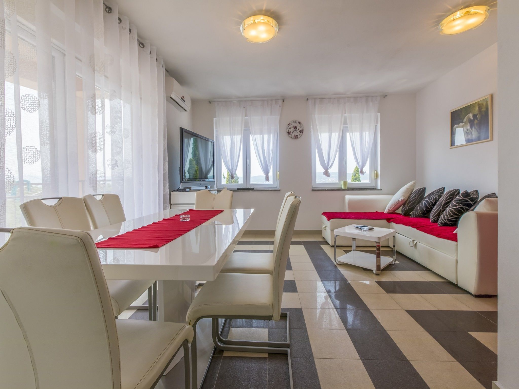 Un salon moderne comprenant un coin repas, des meubles blancs, un téléviseur à écran plat et de grandes fenêtres ornées de rideaux légers.