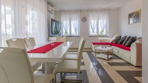 Ein modernes Wohnzimmer mit Essbereich, weißen Möbeln, einem Flachbildfernseher und großen Fenstern mit transparenten Vorhängen.