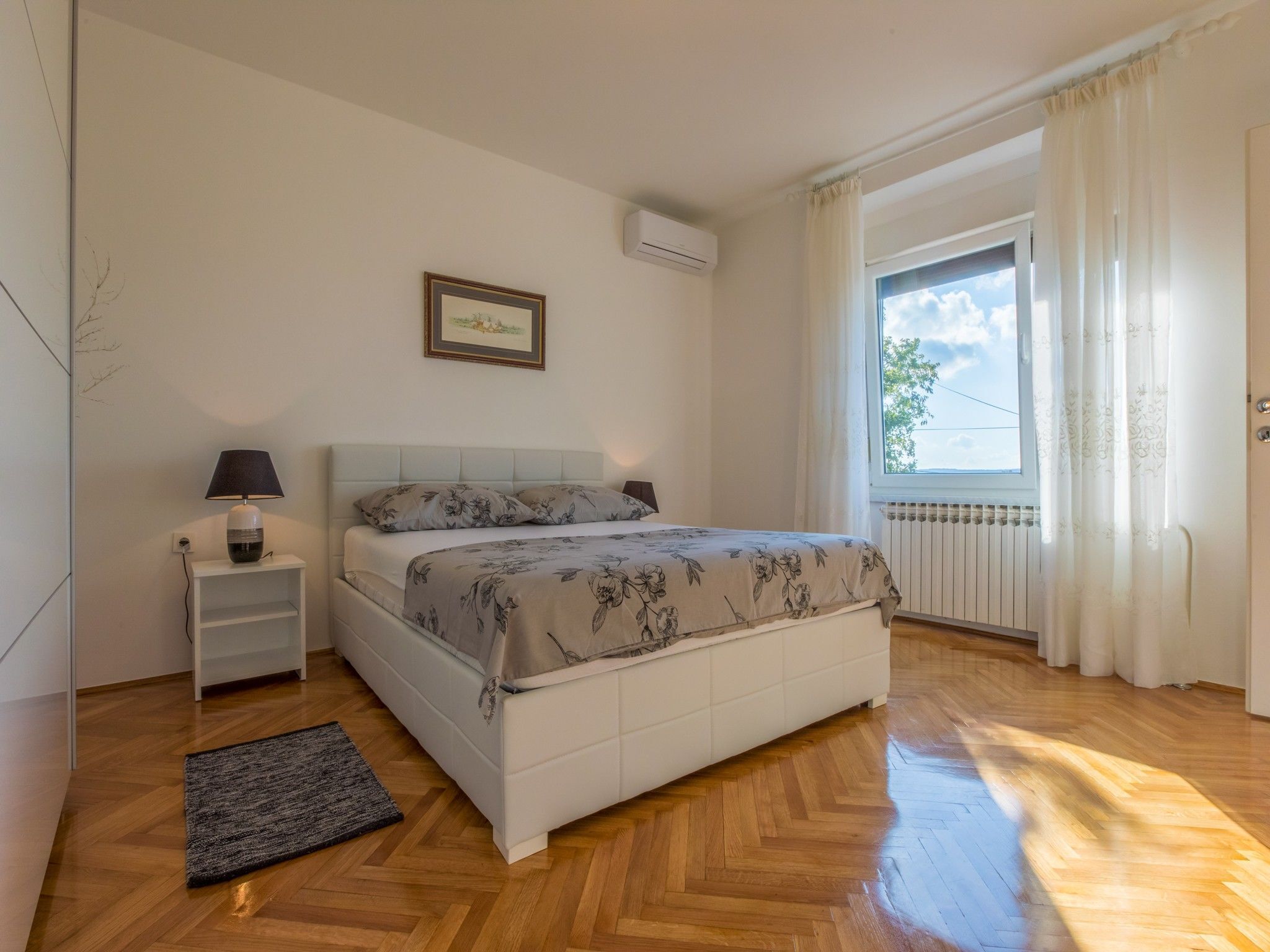 Een slaapkamer met een bed, een nachtkastje met een lamp, een radiator, een raam met gordijnen, en een lijst met een afbeelding aan de muur.