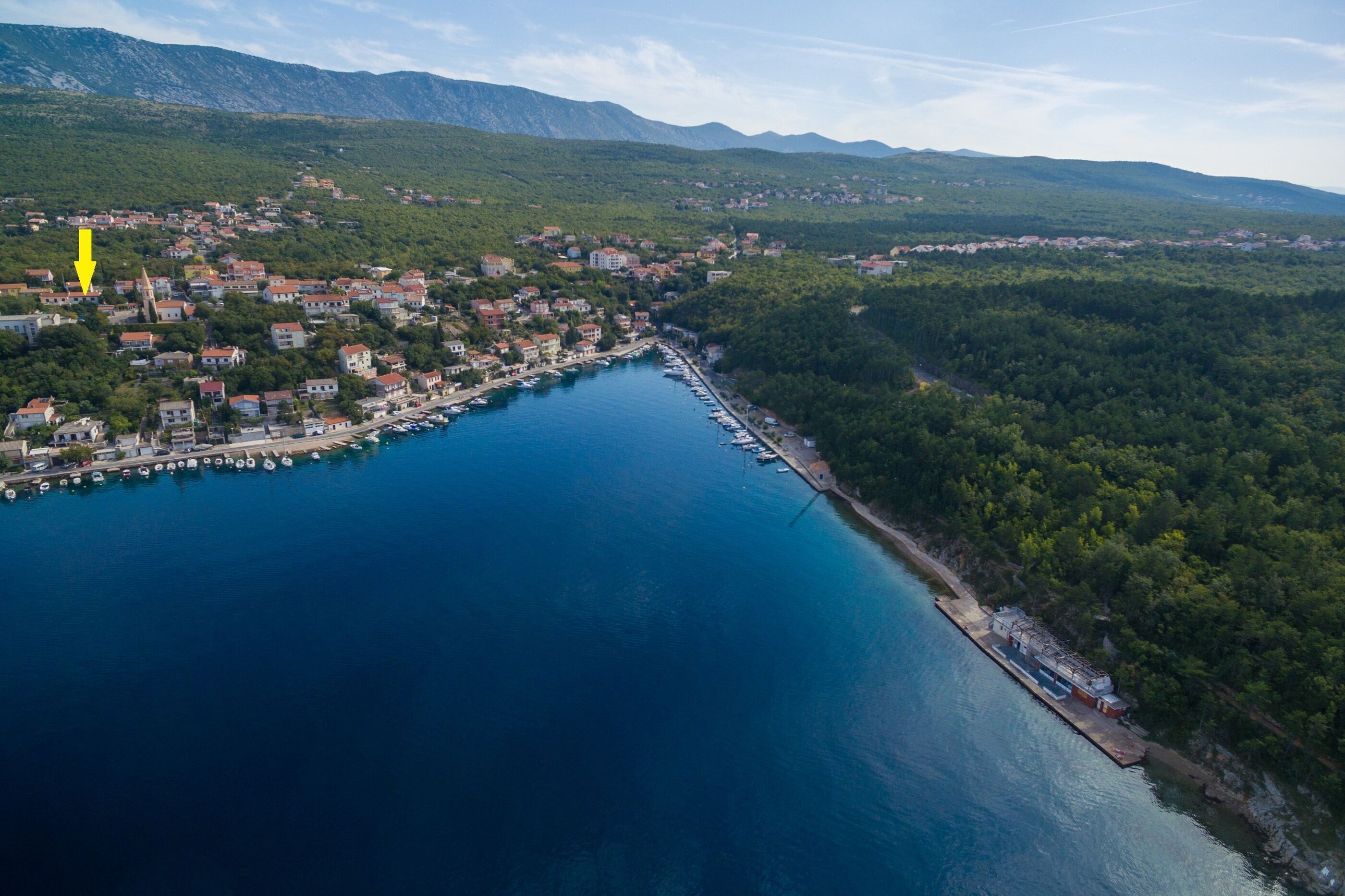 An aerial view of a coastal town with a marina, surrounded by green hills and mountains.