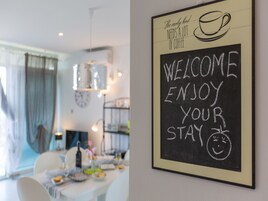 A dining area with a table set for two, a coffee cup illustration, and a chalkboard sign that reads "WELCOME ENJOY YOUR STAY".