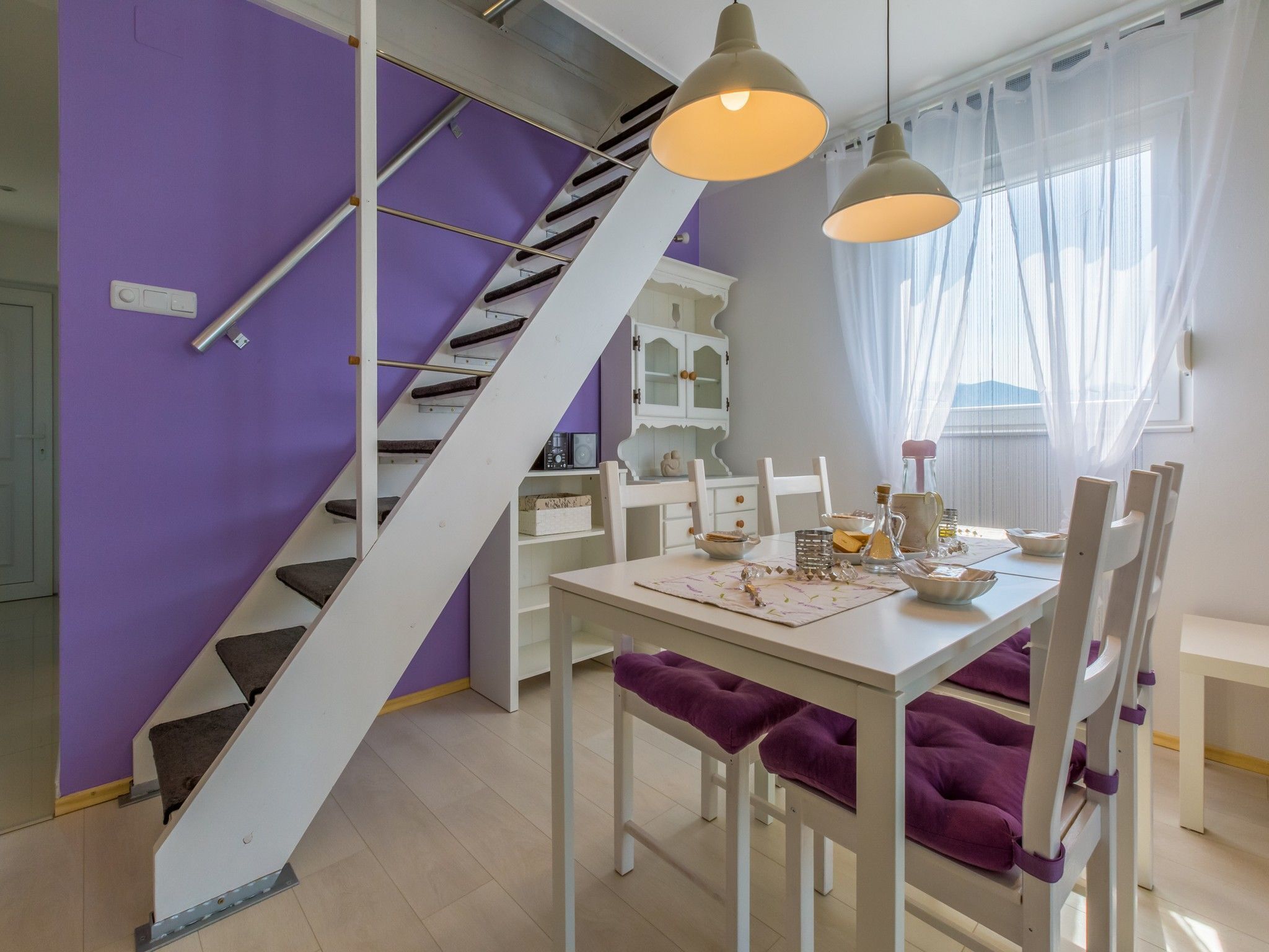 Un espace repas avec une table blanche, des chaises à coussins violets, un escalier blanc et des luminaires suspendus.
