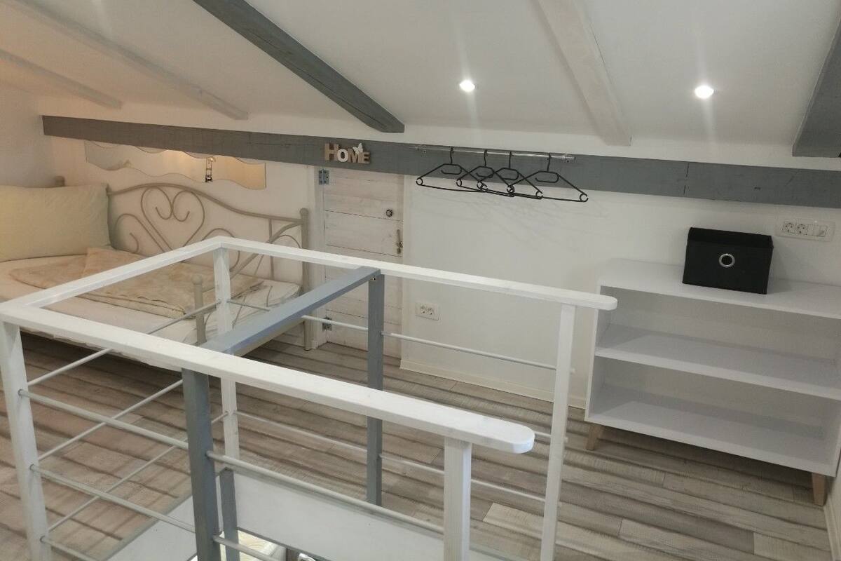 A room with a bed, a white metal crib, a white shelf unit, and a 'HOME' sign on the wall.